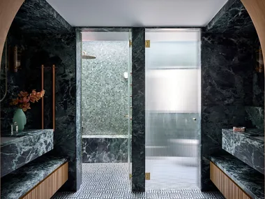 Luxurious bathroom with green marble, wood accents, mosaic tiles, and glass shower doors leading to separate shower and toilet areas. An arch is in the foreground, leading from the bedroom to the bathroom. The room is symmetrical beyond that.