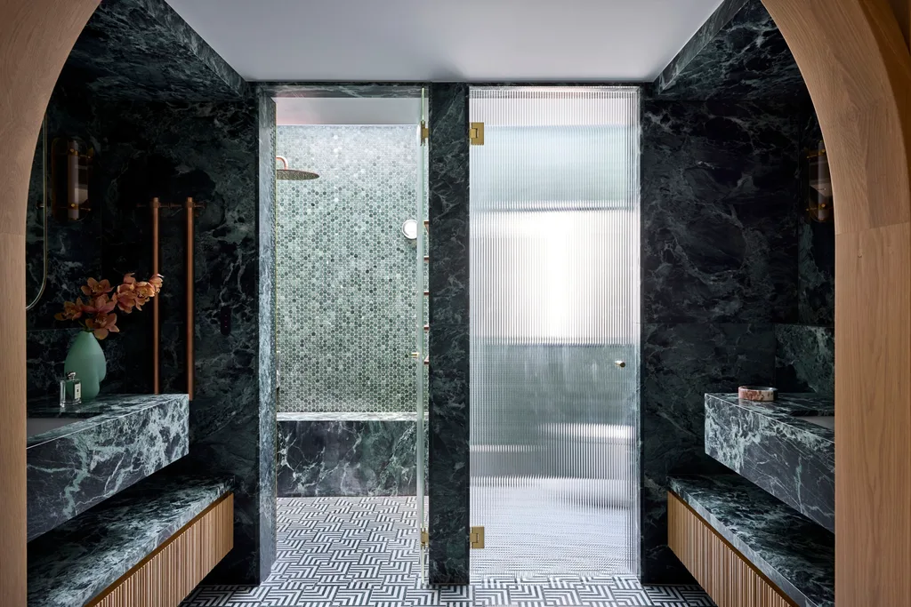 Luxurious bathroom with green marble, wood accents, mosaic tiles, and glass shower doors leading to separate shower and toilet areas. An arch is in the foreground, leading from the bedroom to the bathroom. The room is symmetrical beyond that.