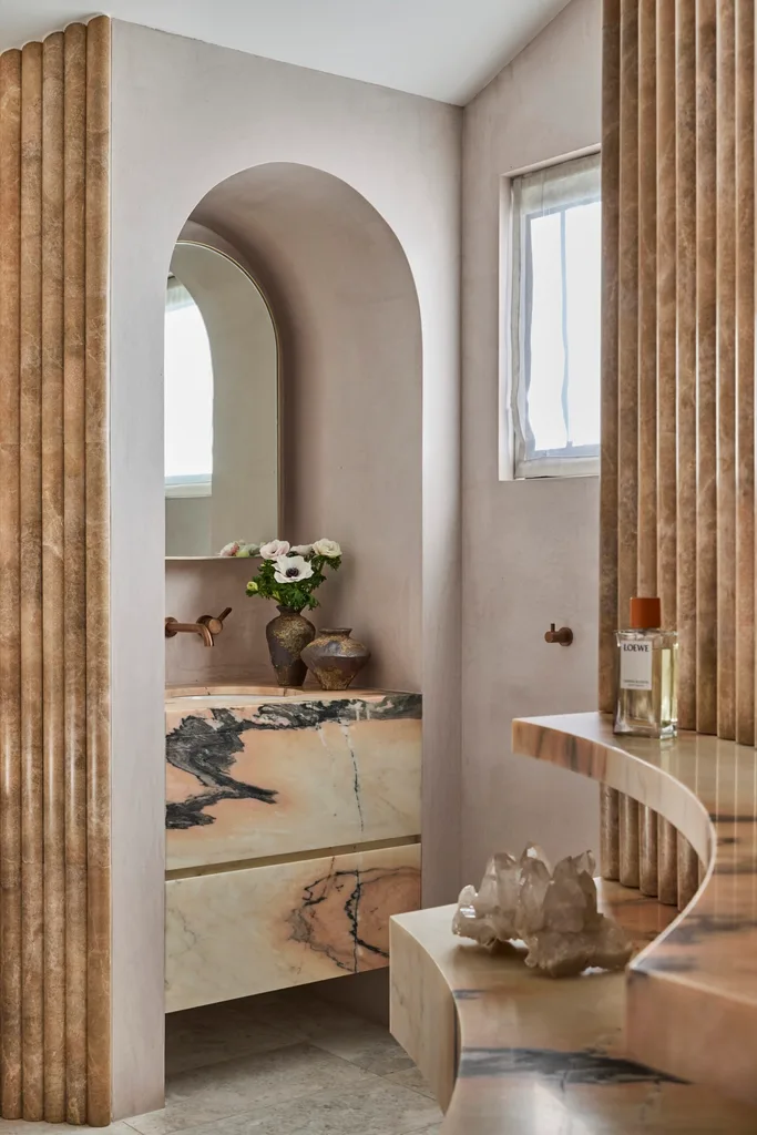 Luxurious bathroom with marble vanity and arched mirror, built in to a large arched alcove. In the foreground, a curved marble shelf has a large crystal and a perfume bottle on it.