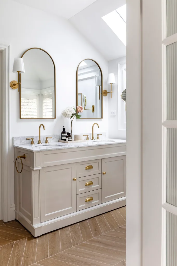 Elegant bathroom with double vanity, marble countertop, gold tapware, two arched mirrors, and wooden herringbone floor.