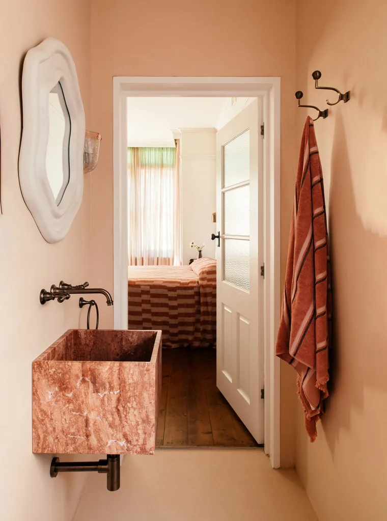 Small ensuite bathroom with pink colour drenched wall and flooring, marble sink, aged brass tapware, and an open white door leading to a bedroom with wooden floorboards and a pink striped bed.