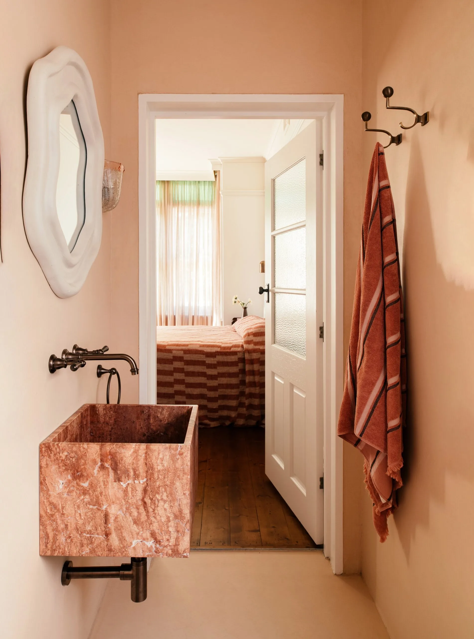 Small ensuite bathroom with pink colour drenched wall and flooring, marble sink, aged brass tapware, and an open white door leading to a bedroom with wooden floorboards and a pink striped bed.