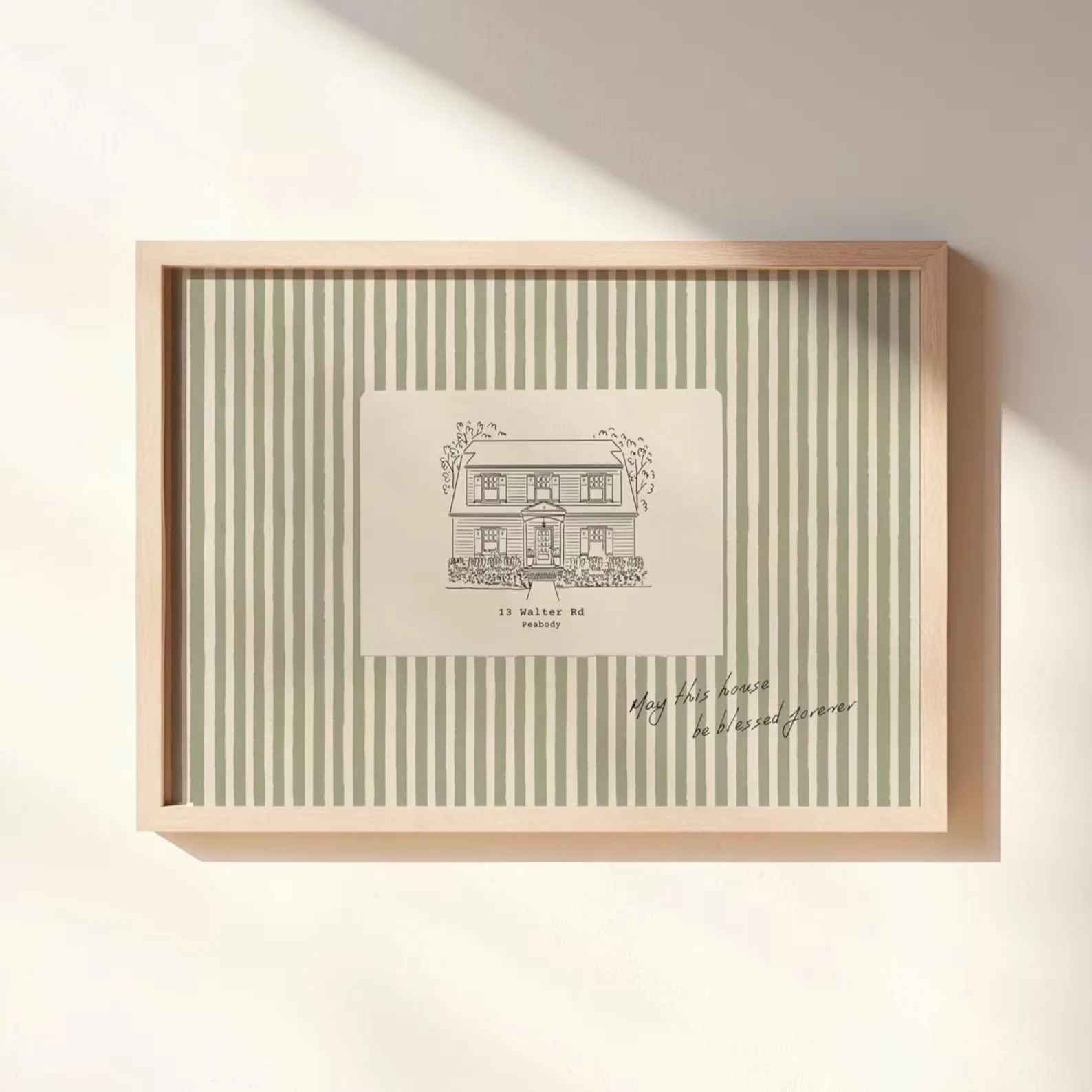 A framed drawing of a home, with the address printed underneath. The frame is a light toned wood and the backboard is striped green and cream.