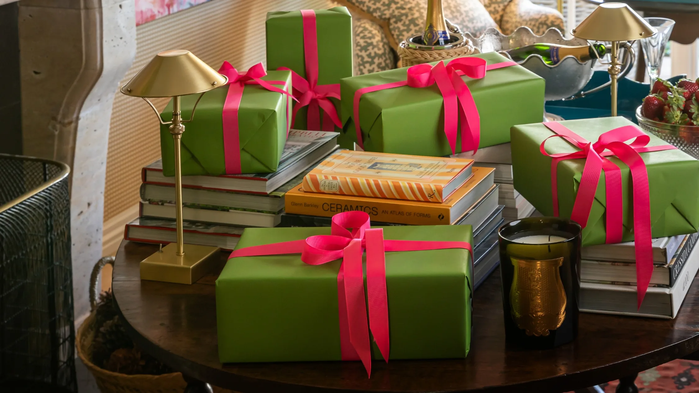 A pile of pretty and neatly wrapped presents on a round dining table covered in coffee table books, golden lamps and a candle. The wrapping paper is green and each gift has a bright pink bow tied on it.