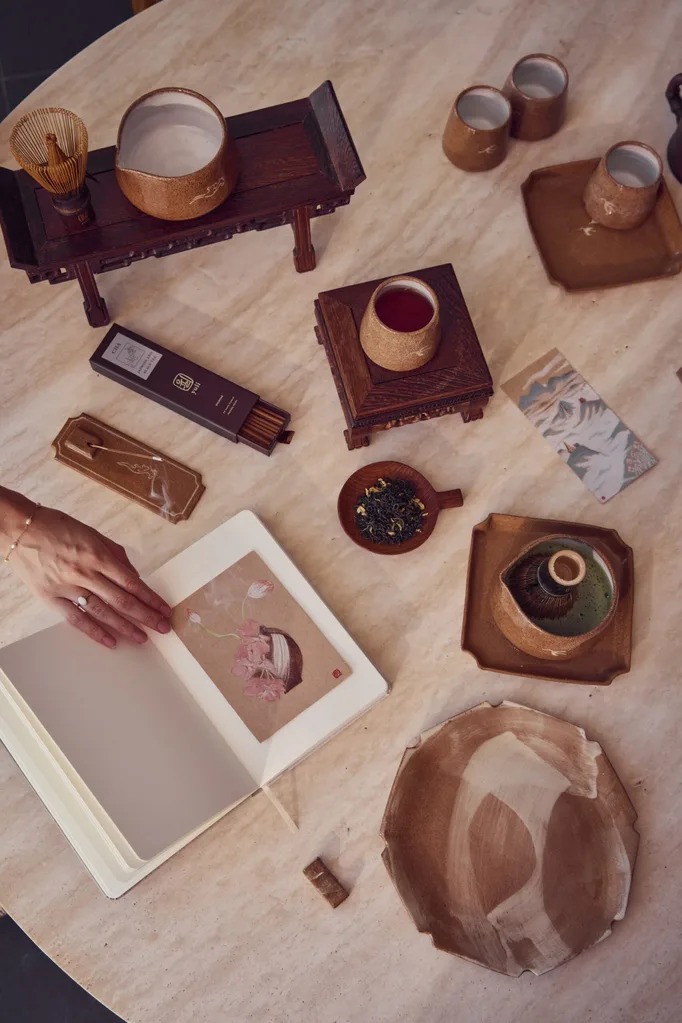 A photo taken from above of a round, stone table with various ceramics on it. The stoneware includes small jugs, mugs and plates. A notebook is open with a small painting on a moon vase with flowers inside.