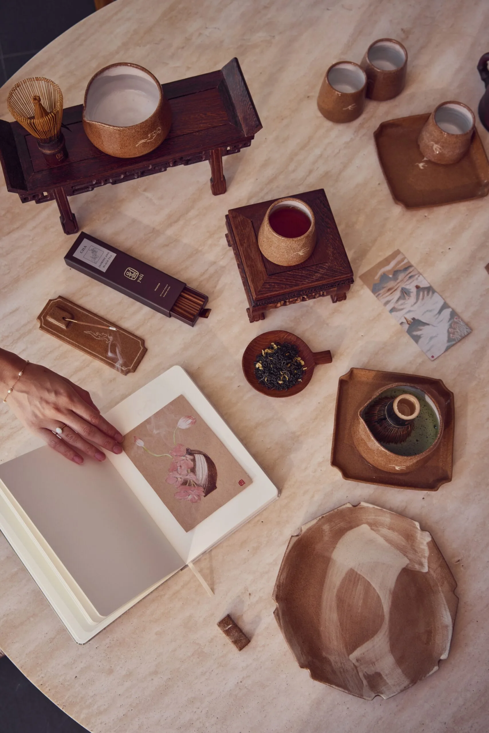 A photo taken from above of a round, stone table with various ceramics on it. The stoneware includes small jugs, mugs and plates. A notebook is open with a small painting on a moon vase with flowers inside.