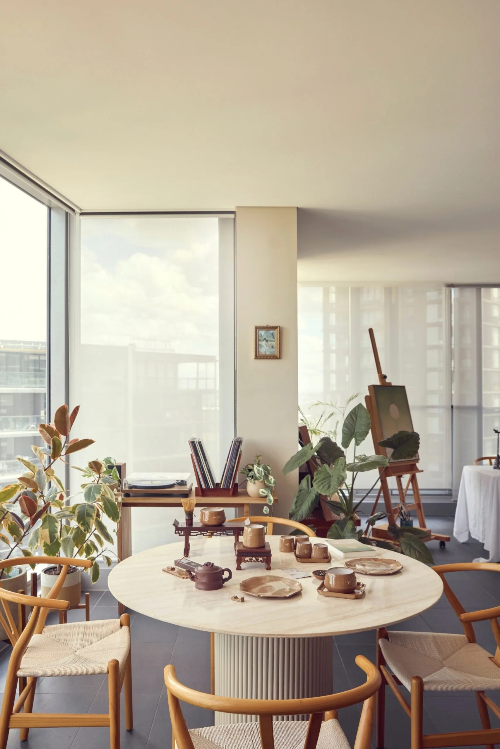 The interior of a high rise apartment. A round stone dining table has four timber dining chairs positioned around it. The room is light and bright, with a small painting in a frame on the wall in the background and a wooden easel nearby it, with a large painting on it. There are also lots of leafy potted plants. The table has handmade earthenware pottery on it, including plates, jugs, mugs and trays.