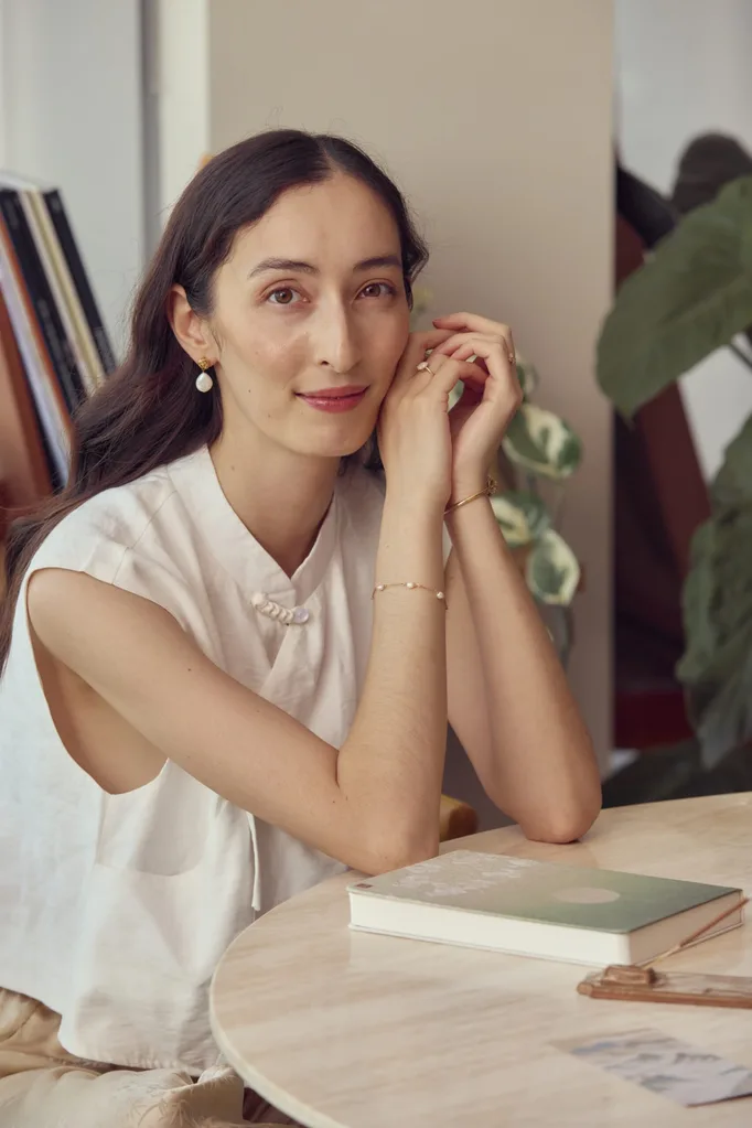 A woman in a sleeveless white shirt sits at a round dining table. Her face rests on her hands while her elbows rest on the table. A green notebook and a stone incense holder are positioned in front of her. She smiles softly.