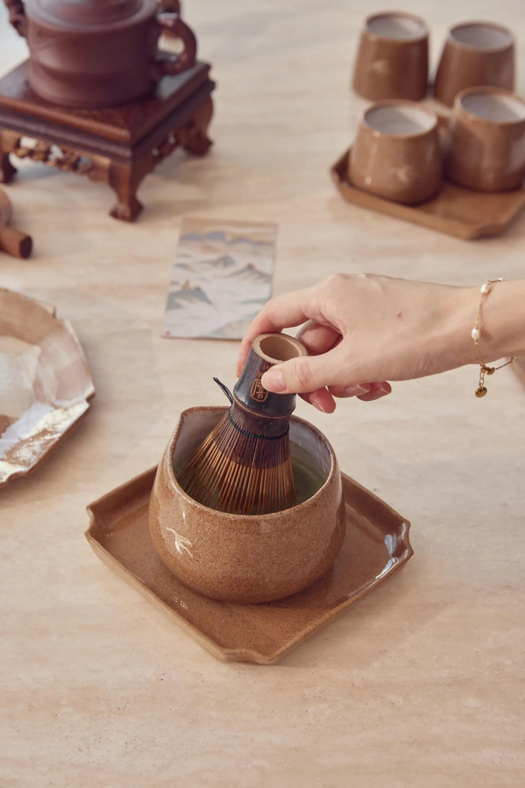 A close up photo of handmade stoneware mugs and trays. A hang is holding a matcha whisk, making matcha in one of the mugs.