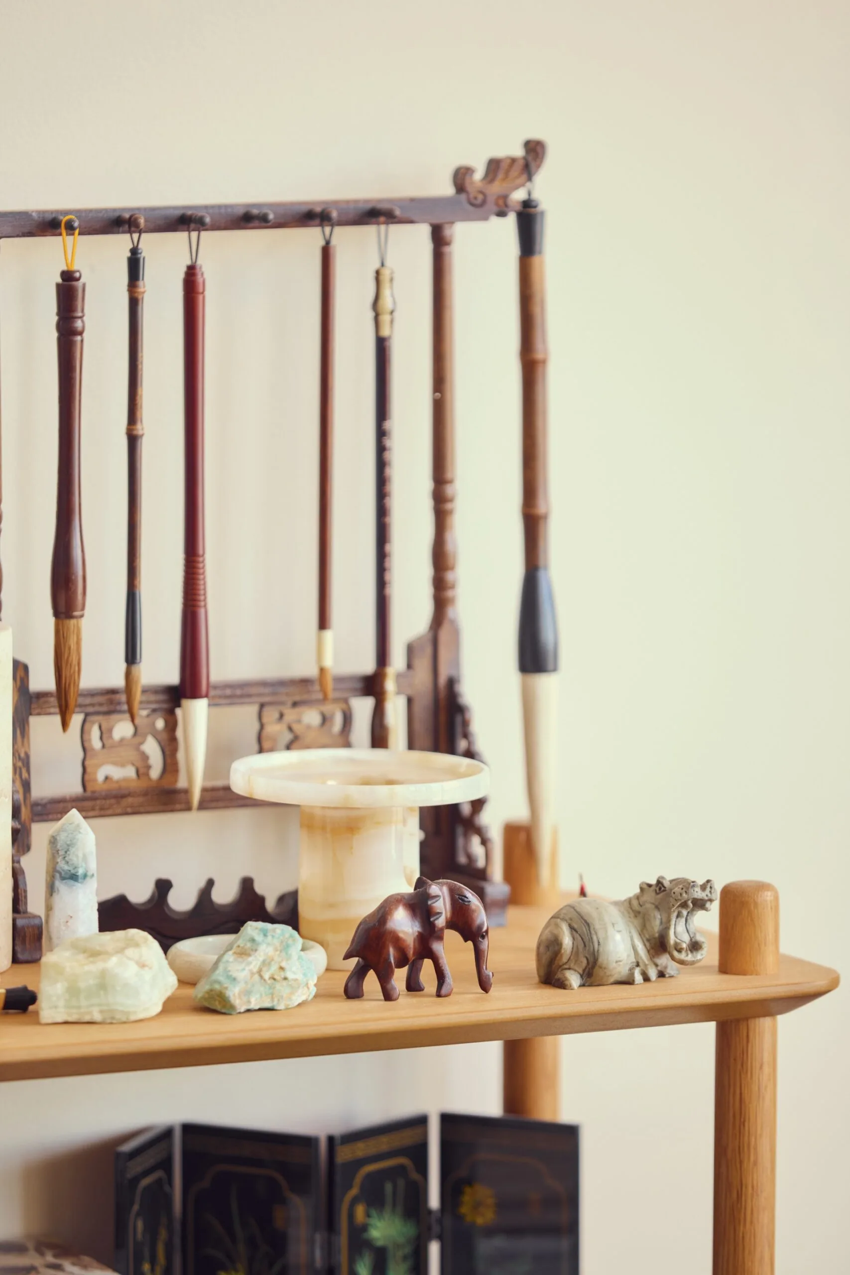 A close up photo of a console table. A little timber frame has hooks across the top that paintbrushes hang from. Jade stamps, crystals and small animal figurines are displayed in front of this.