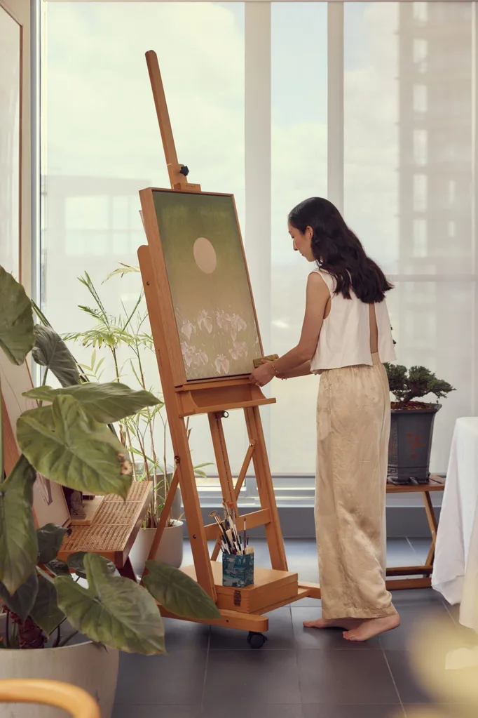 A woman in cream toned pants and a white sleeveless shirt stands in front of a wooden easel. She is using a jade stamp to put her initials on the painting, which is a calming green colour and depicts the moon above a field of irises.
