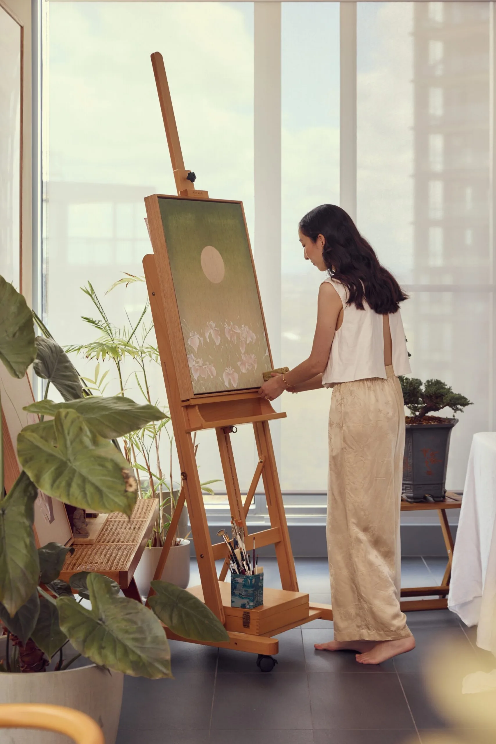 A woman in cream toned pants and a white sleeveless shirt stands in front of a wooden easel. She is using a jade stamp to put her initials on the painting, which is a calming green colour and depicts the moon above a field of irises.