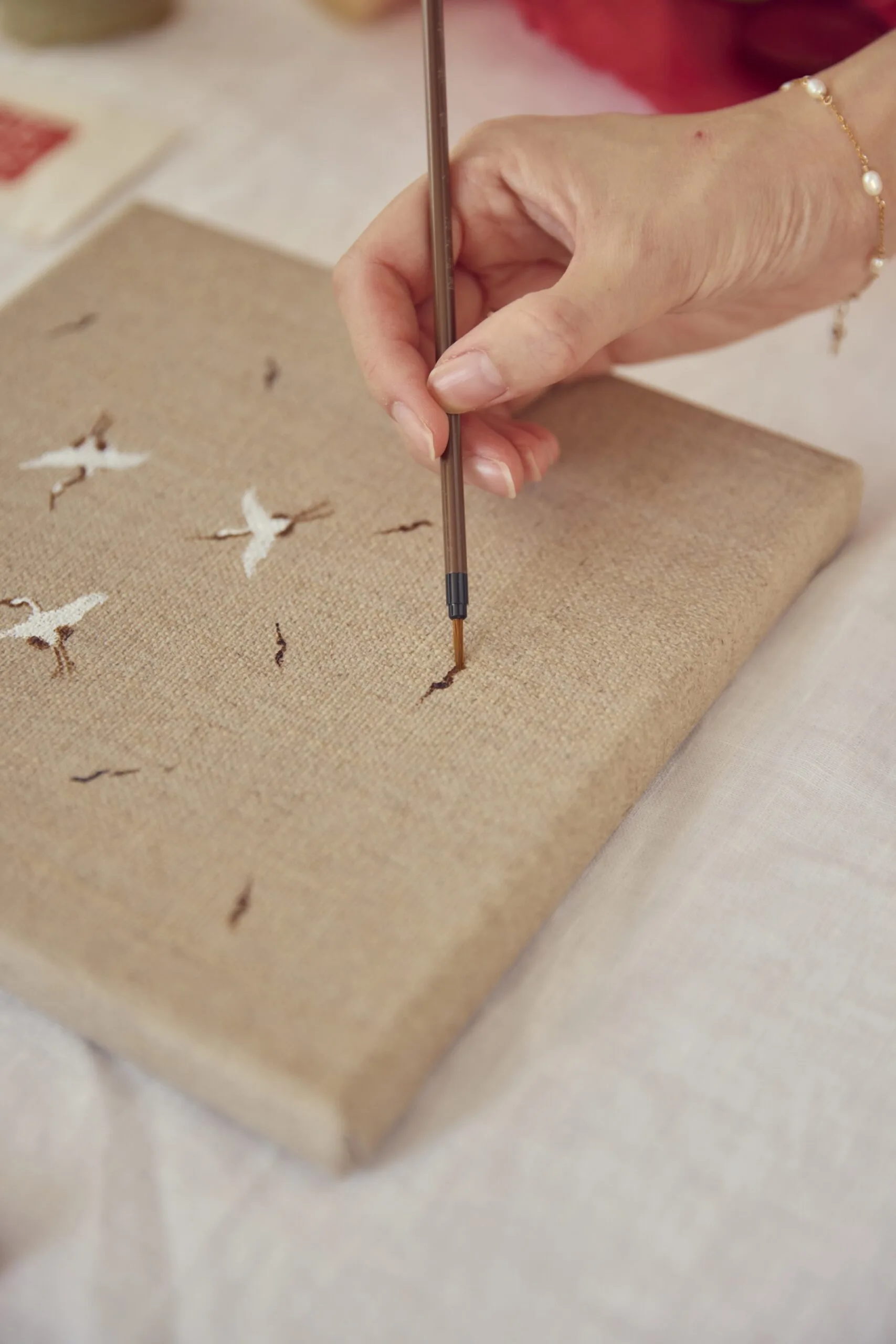 A close up photo of a hand holding a small paintbrush. The person is painting a small image of cranes on to a brown toned canvas.