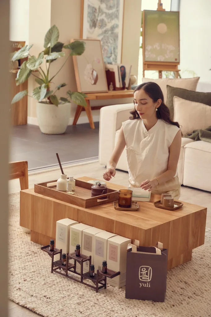 A woman sits on a cream coloured rug, in front of a wooden coffee table. There is a cream coloured rug in the background and three large paintings. The coffee table has incense, reed diffusers and candles on it, all from Yuli Kiln. A shopping bag is in front of the coffee table with Yuli Kiln's logo on it.