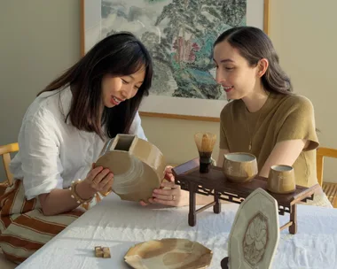 Two women sit at a rectangular dining table with a white tablecloth on top. They are looking down and smiling, holding a handmade earthenware moon vase. Other handmade ceramic items, including plates and jugs, are positioned on the table in front of them. A large painting of a landscape by a Chinese artist is on the wall behind them.