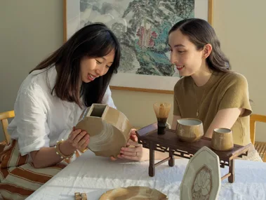Two women sit at a rectangular dining table with a white tablecloth on top. They are looking down and smiling, holding a handmade earthenware moon vase. Other handmade ceramic items, including plates and jugs, are positioned on the table in front of them. A large painting of a landscape by a Chinese artist is on the wall behind them.