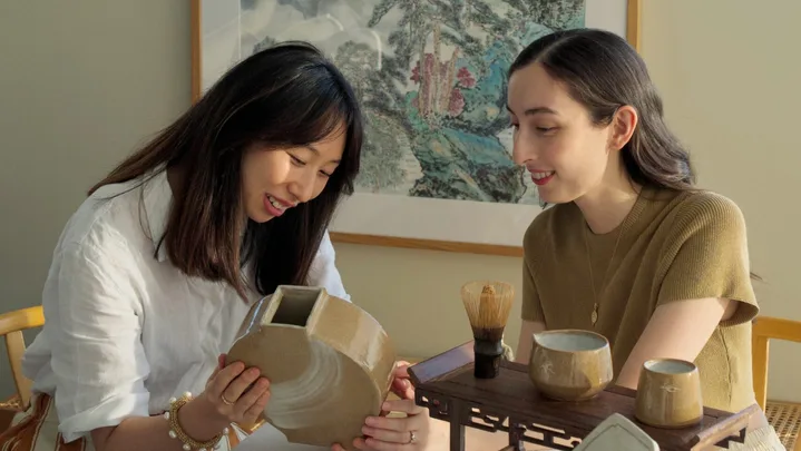 Two women sit at a rectangular dining table with a white tablecloth on top. They are looking down and smiling, holding a handmade earthenware moon vase. Other handmade ceramic items, including plates and jugs, are positioned on the table in front of them. A large painting of a landscape by a Chinese artist is on the wall behind them.