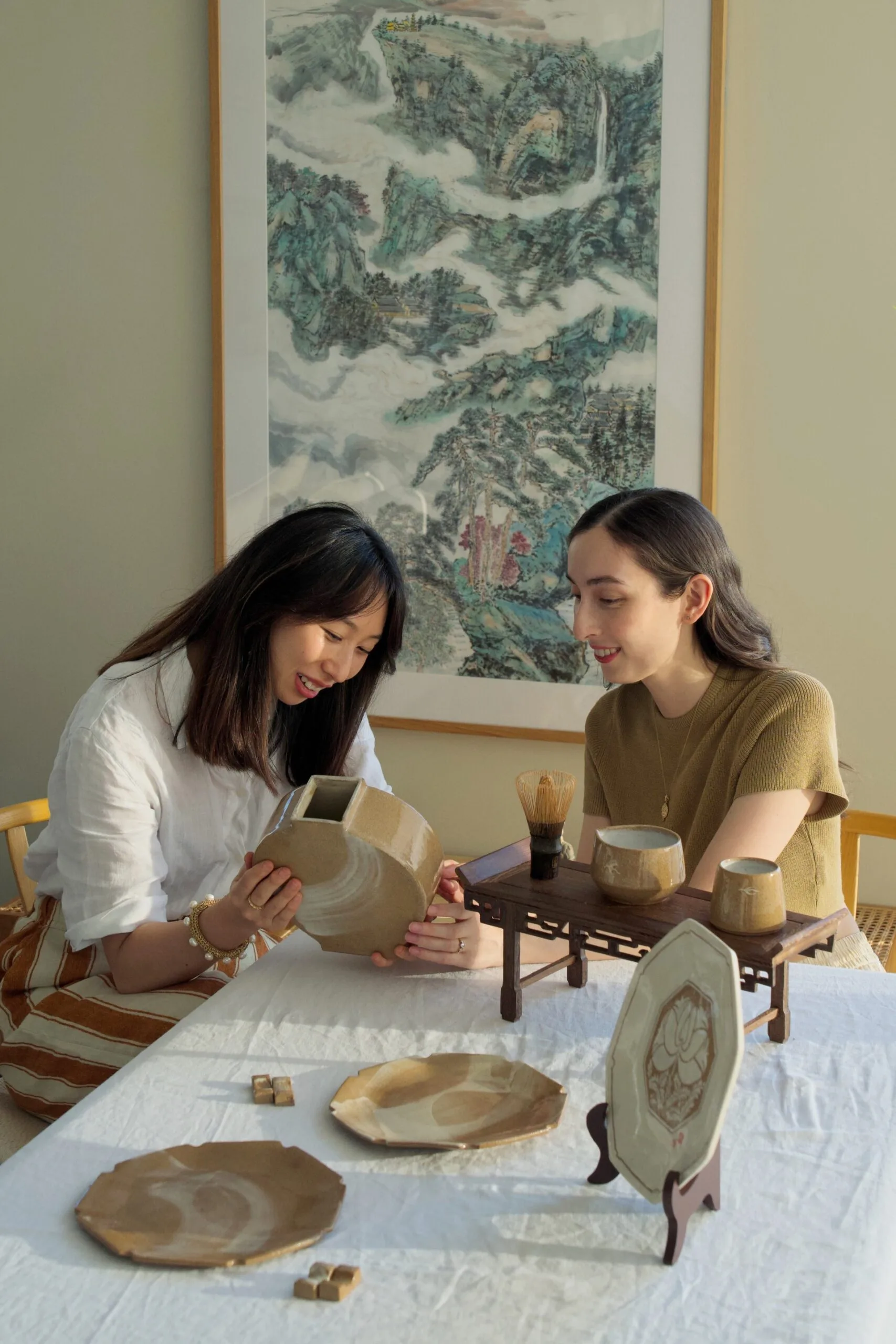 Two women sit at a rectangular dining table with a white tablecloth on top. They are looking down and smiling, holding a handmade earthenware moon vase. Other handmade ceramic items, including plates and jugs, are positioned on the table in front of them. A large painting of a landscape by a Chinese artist is on the wall behind them.