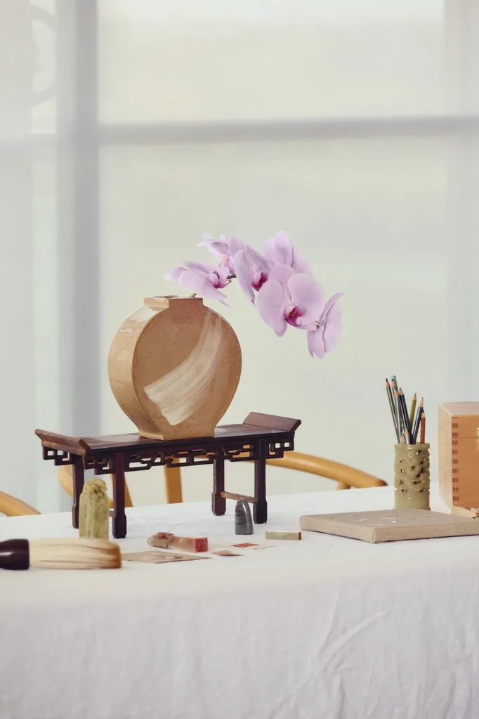 A handmade moon vase has one stalk of pink orchids displayed in it. This is positioned on a table with a white tablecloth, beside jade stamps, a small canvas, and a jade pencil holder with watercolour pencils in it.
