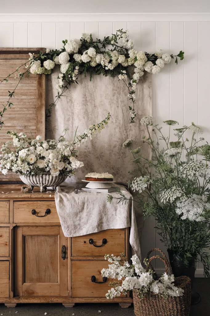 Vintage wooden cabinet with lush white floral arrangements, a cake on a stand, and a basket of white blooms beside.