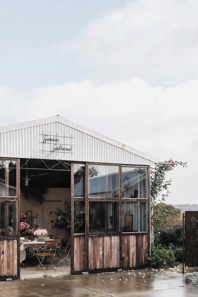 Exterior of a rustic floristry shed with open doors, surrounded by greenery and pink flowers, under a cloudy sky.
