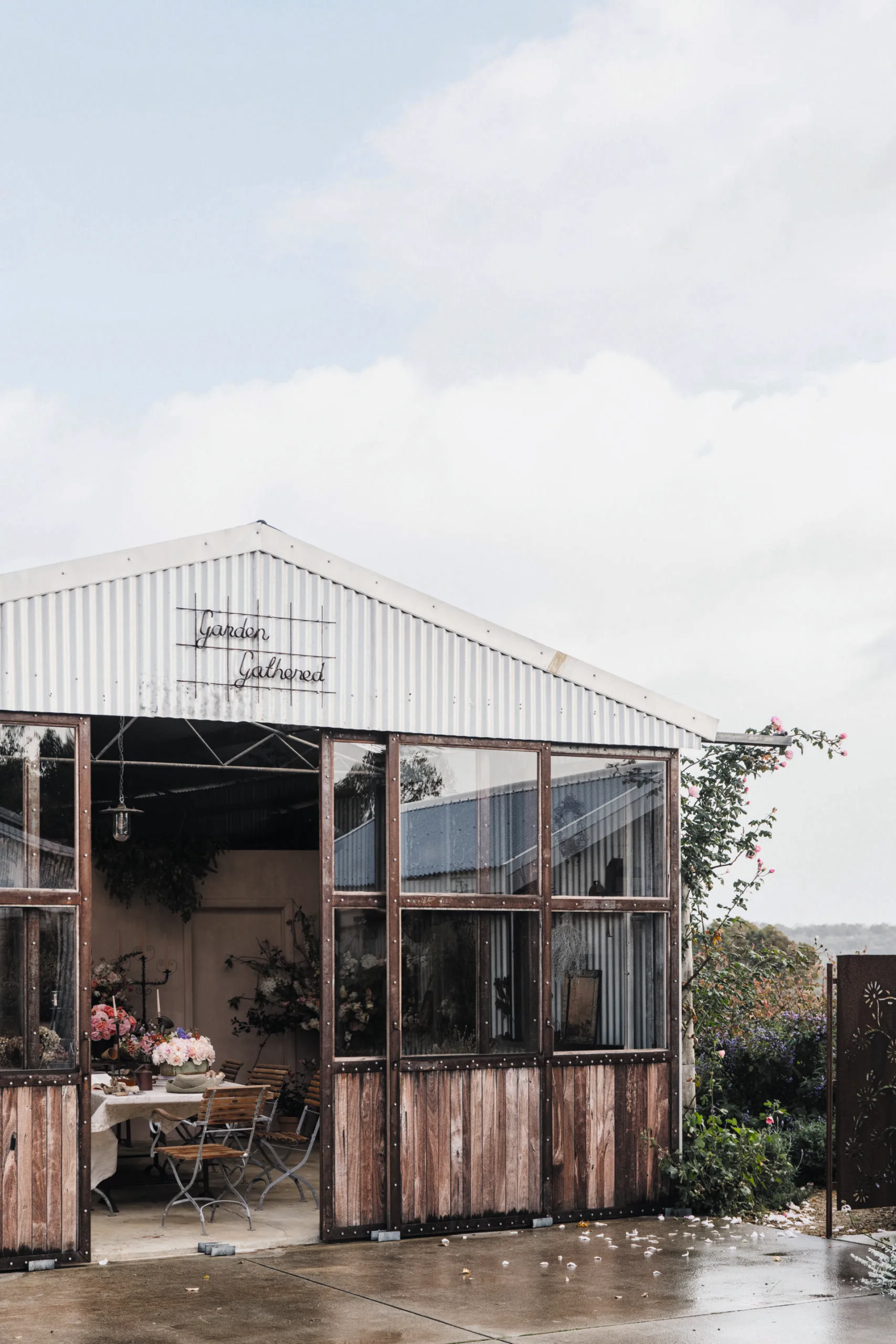 Exterior of a rustic floristry shed with open doors, surrounded by greenery and pink flowers, under a cloudy sky.