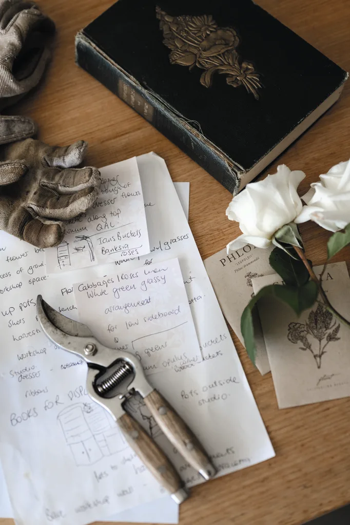 Gardening notes and pruning shears on wooden table with a book, gloves, and white roses.