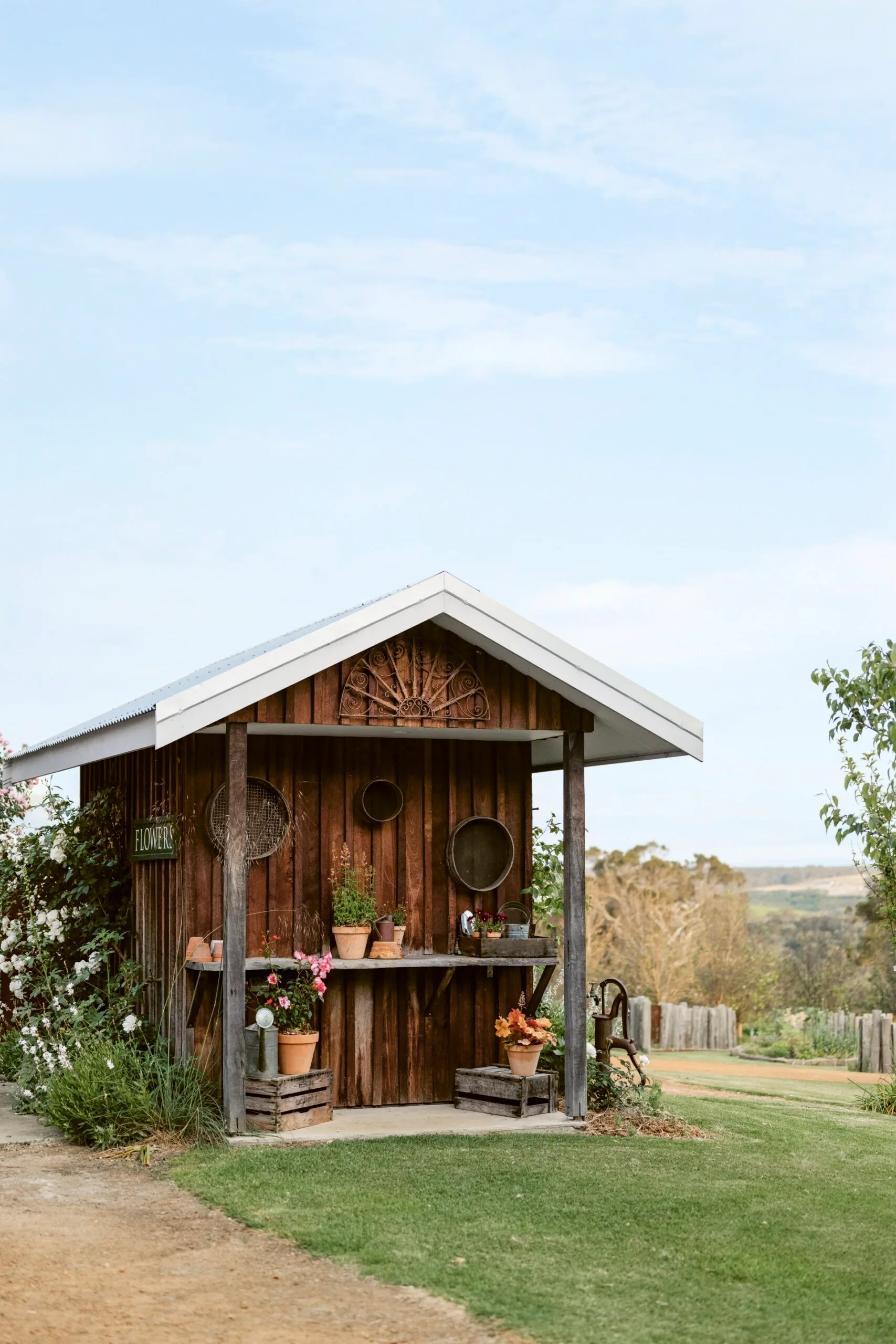 Rustic wooden flower shed with potted plants and vintage decor, set against a clear blue sky and rural landscape.