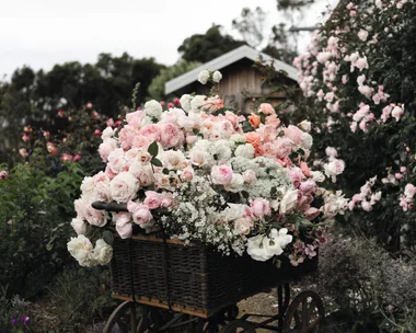 A vintage cart or wheelbarrow filled with pink and white roses, as well as other pink and white flowers, flowers in a lush garden setting.