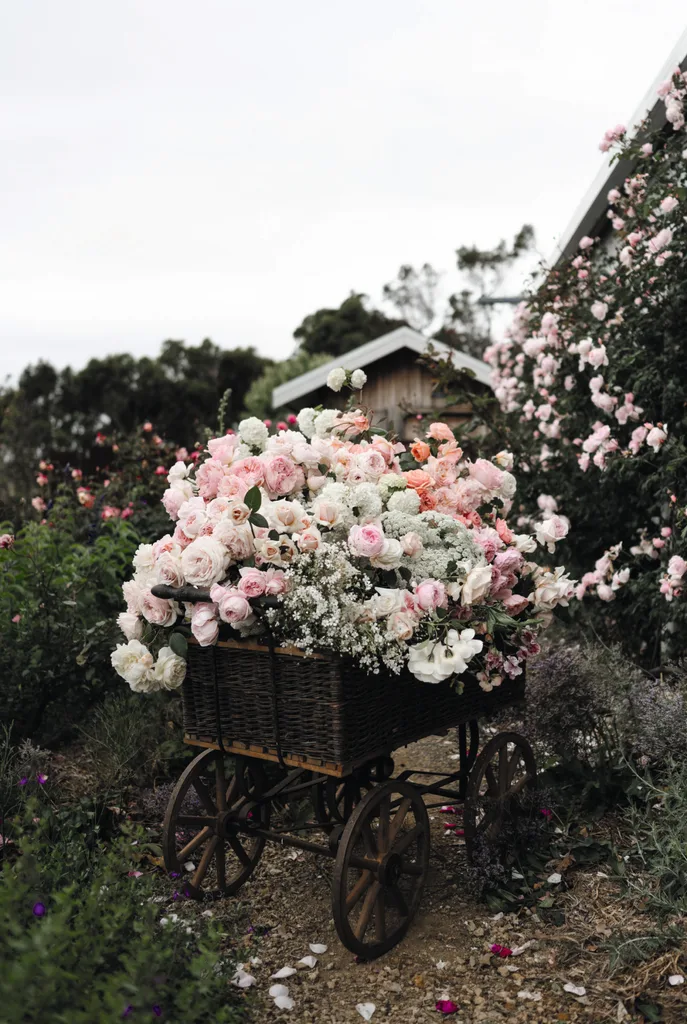 A vintage cart or wheelbarrow filled with pink and white roses, as well as other pink and white flowers, flowers in a lush garden setting.