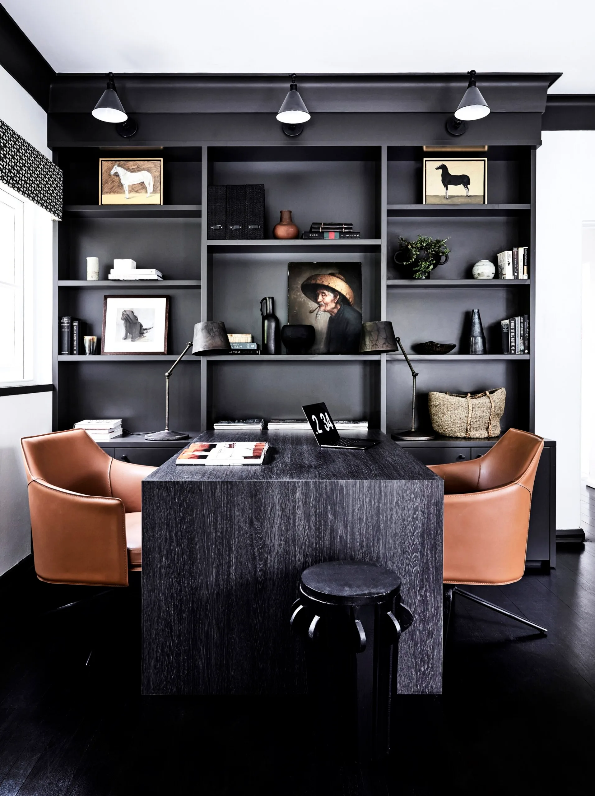 Sleek modern home office with dark wood desk, leather chairs, and art-filled shelves. The open shelves are finished in a black shade, matching the desk.