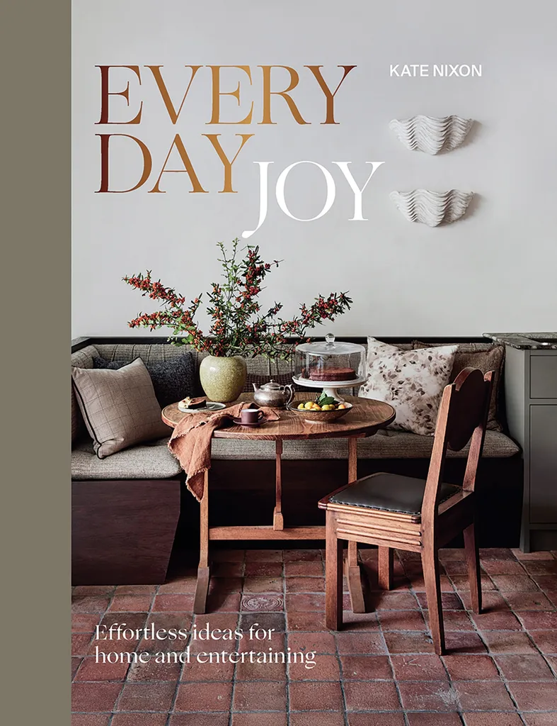 Cover of "Everyday Joy" by Kate Nixon, featuring a dining area with timber round dining table, one dining chair, an L-shaped banquette with grey upholstery and decorative items on the table such as a vase with a bouquet and a cake stand.