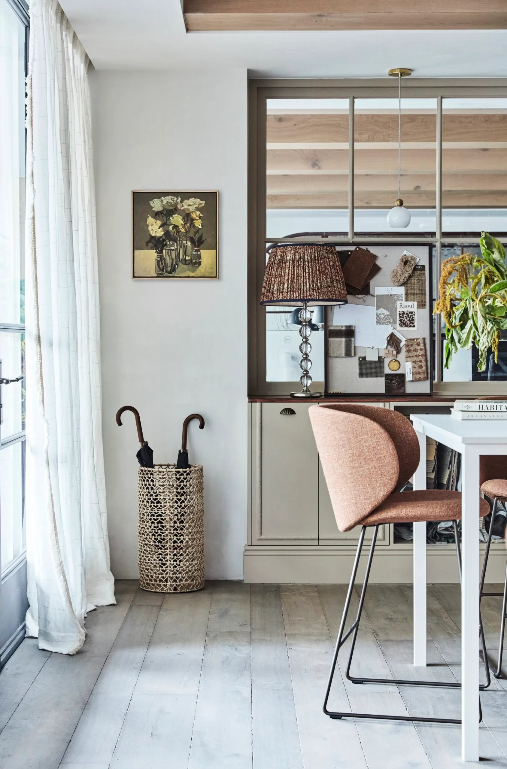 Cozy room with a wicker umbrella stand, wall art, a cushioned bar stool pulled out from a tall white table, and a bulletin board beside a curtained window.