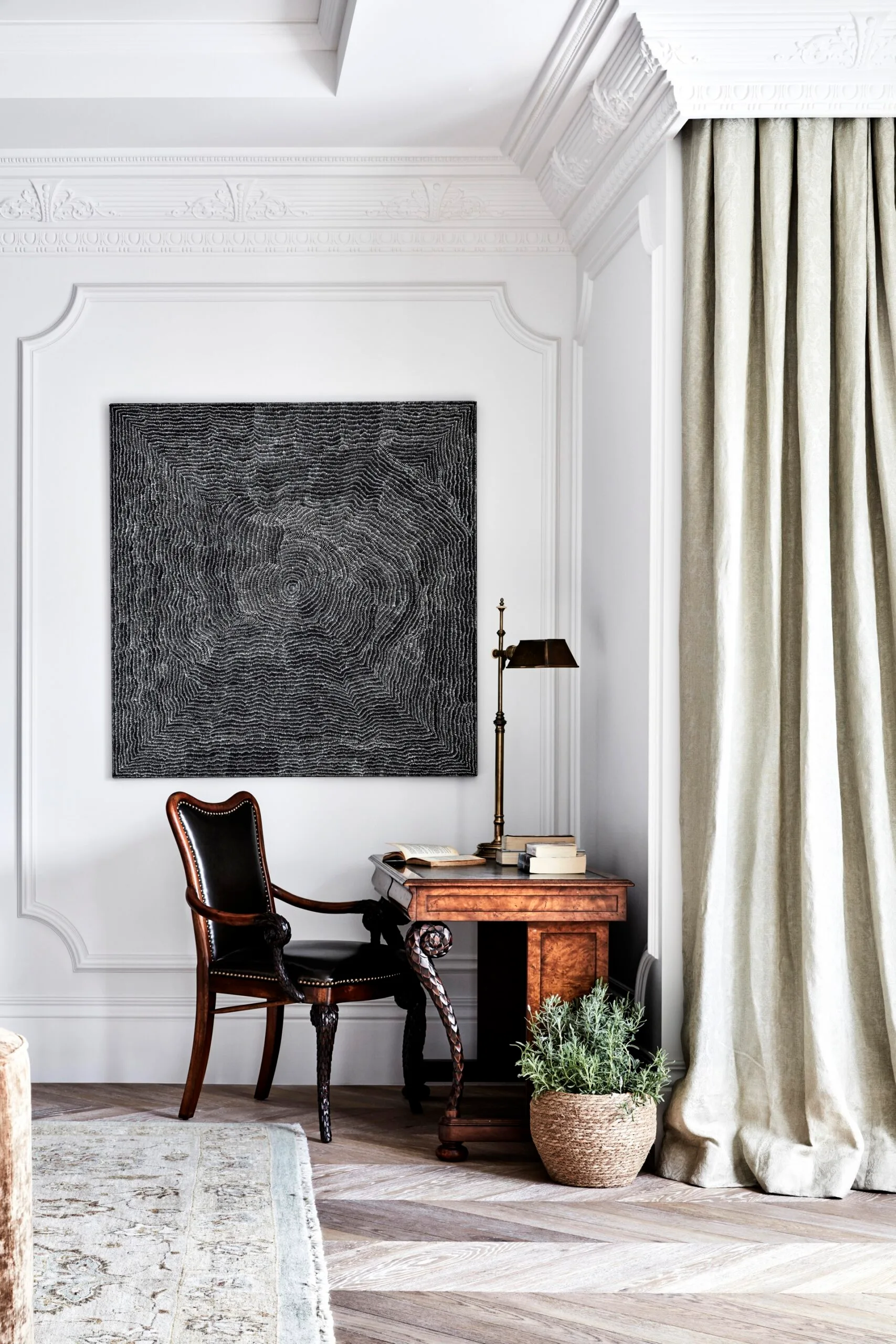 A home office nook with antique wooden desk and chair, table lamp, books, and small potted plant on the floor beside the desk. Large textured artwork on the wall. Elegant and classic decor, including decorative cornices and wall mouldings.