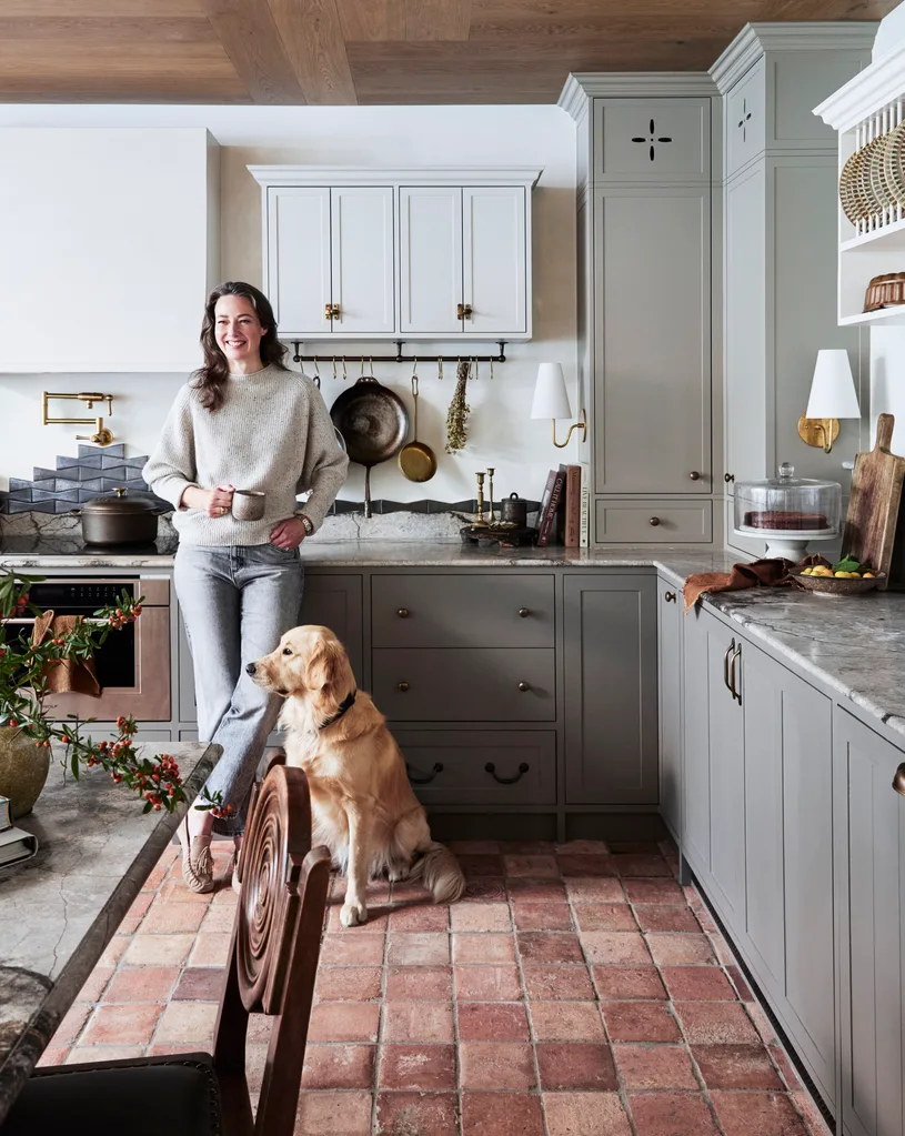 Woman stands in cosy kitchen with a mug, leaning on a marble counter, with a golden retriever sitting beside her. The flooring is tiled, red and rustic while the joinery is finished in a grey shade with shaker-style profile and brass hardware.