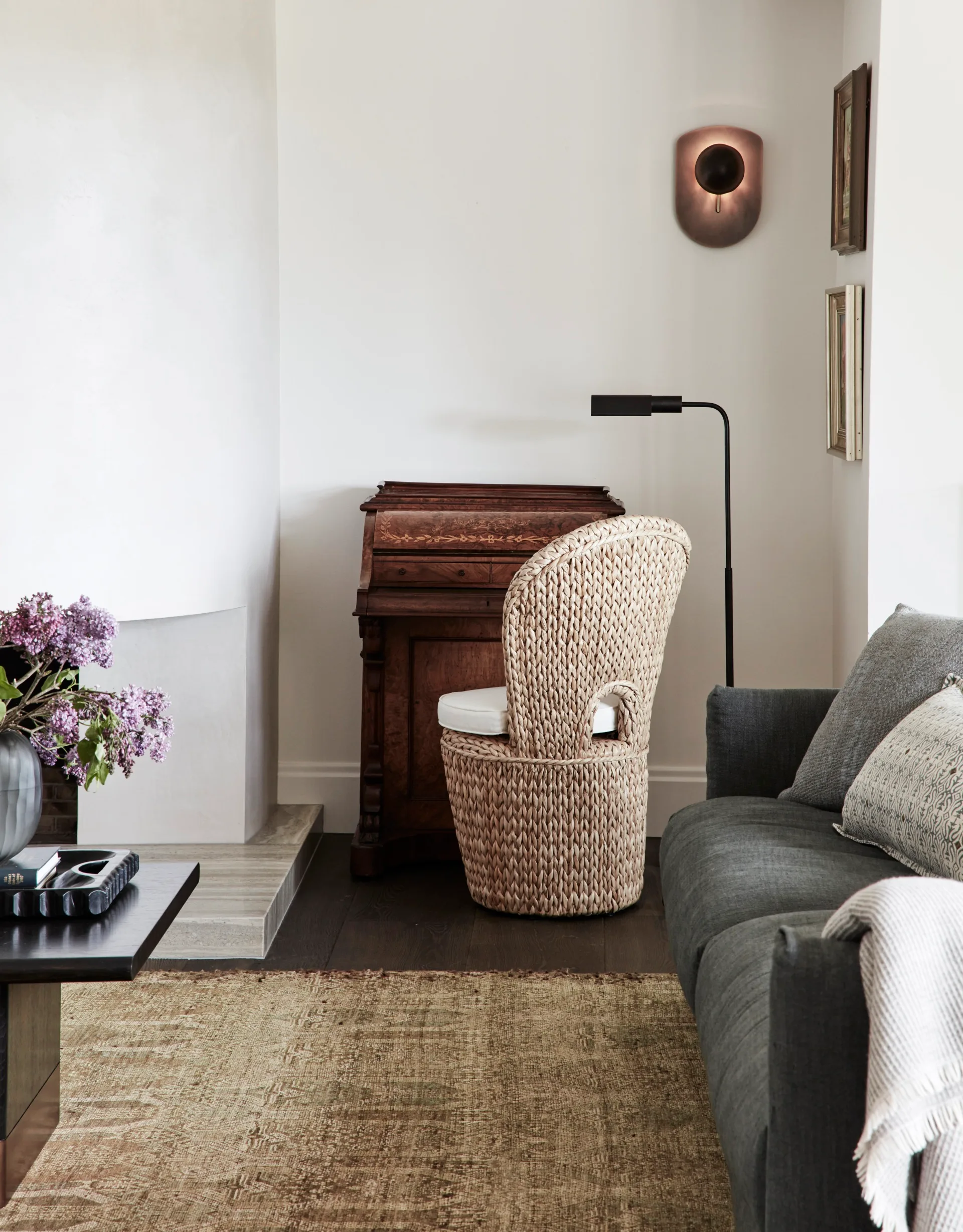 Cozy living room with a home office nook in one corner. This nook includes a wicker chair, wooden desk, and floor lamp. A modern wall sconce is positioned above. In the foreground a gray sofa and coffee table with purple flowers in a vase is visible, as is a neutral toned aged rug.