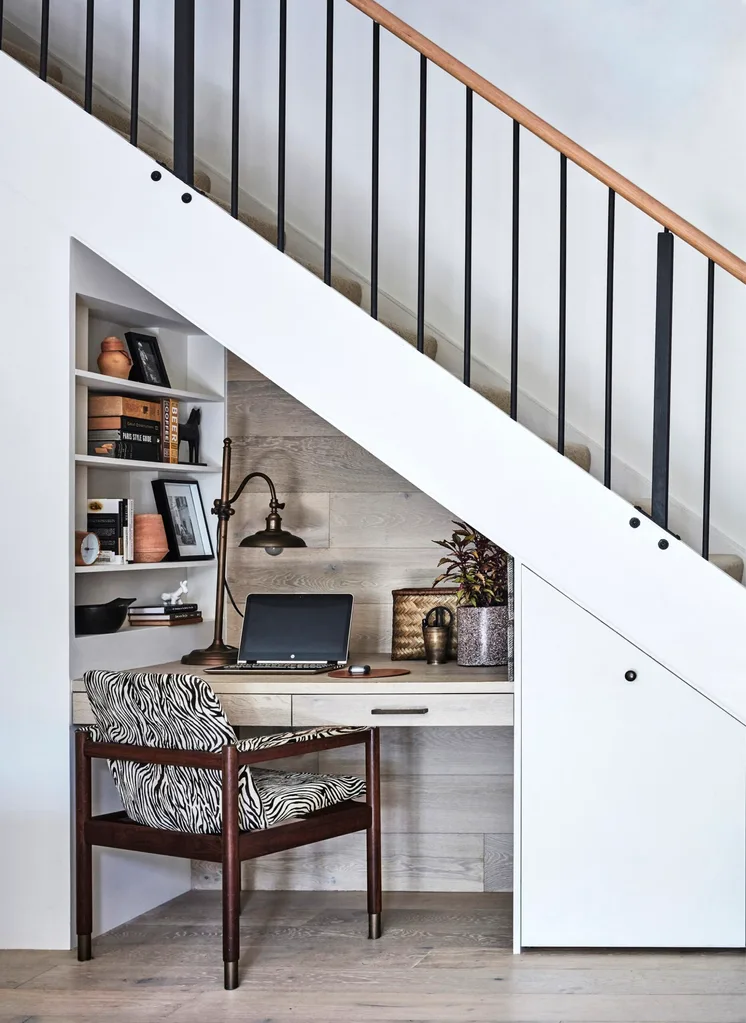 Under-stair workspace with a desk, laptop, table lamp, and zebra-patterned chair. Shelves hold books and decor items. This is all built into the space under the stairs and is open to the rest of the room.