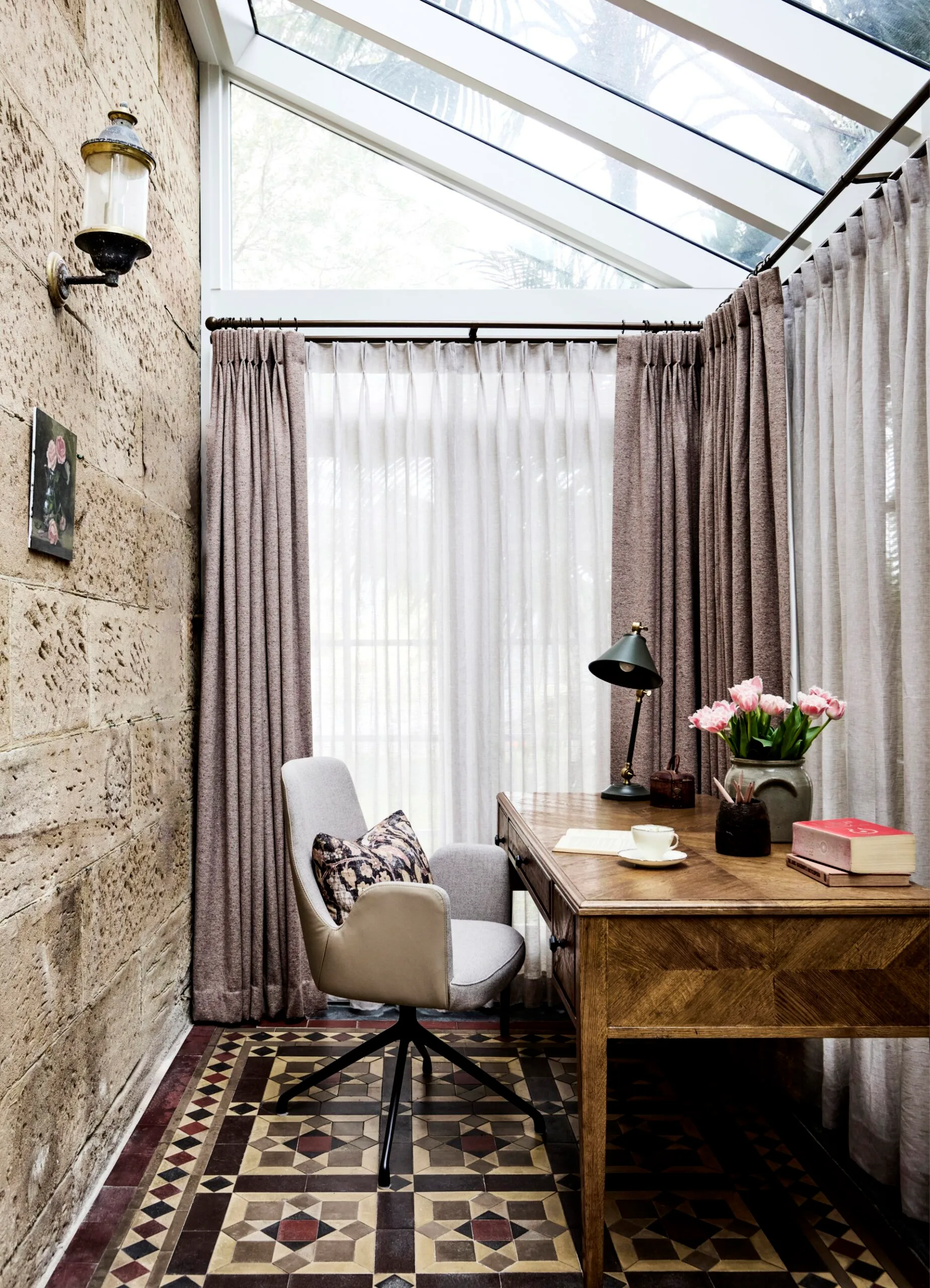 Cozy home office with a wooden desk, lamp, tulips in a vase, and a cushioned desk chair under a vaulted glass ceiling. Two of the walls, underneath the glass ceiling, are also glass with neutral curtains and the wall behind the desk is stone. The floor is patterned tile flooring.