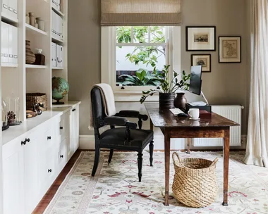 Stylish home office with wooden desk, black desk chair, built-in bookshelf, and window with plants. A sizable rug is on the timber floorboards and art is framed and hung on the wall beside the window.