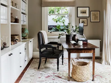 Stylish home office with wooden desk, black desk chair, built-in bookshelf, and window with plants. A sizable rug is on the timber floorboards and art is framed and hung on the wall beside the window.