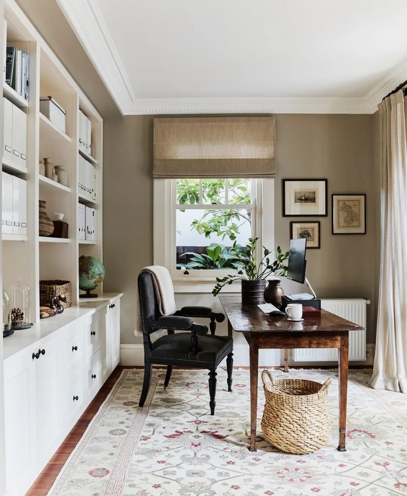 Stylish home office with wooden desk, black desk chair, built-in bookshelf, and window with plants. A sizable rug is on the timber floorboards and art is framed and hung on the wall beside the window.