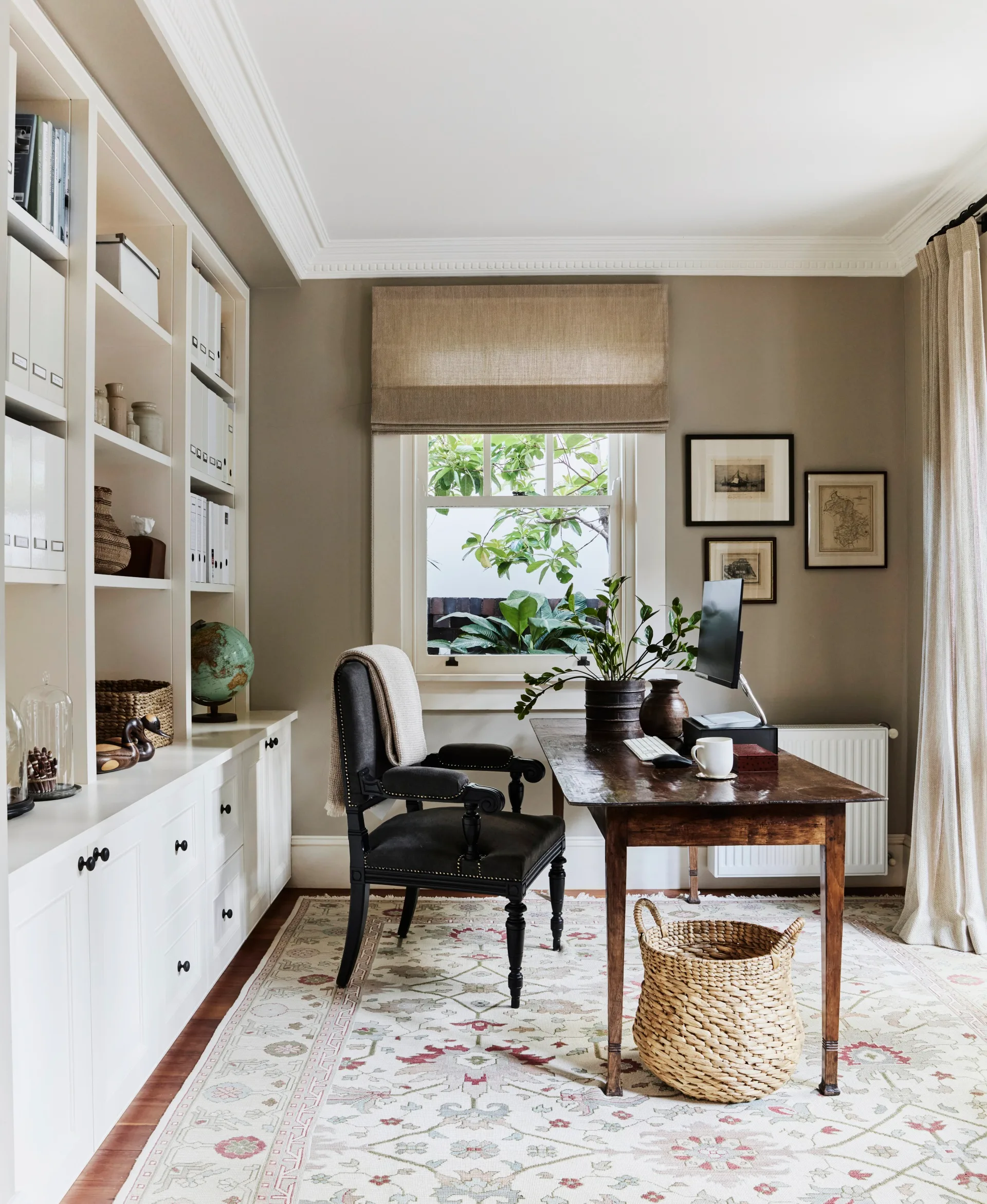 Stylish home office with wooden desk, black desk chair, built-in bookshelf, and window with plants. A sizable rug is on the timber floorboards and art is framed and hung on the wall beside the window.