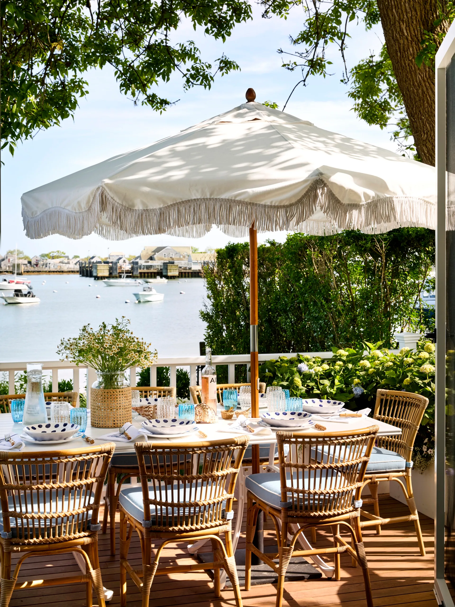 Outdoor dining set with wicker chairs and a white umbrella on a deck, overlooking a calm body of water and boats.