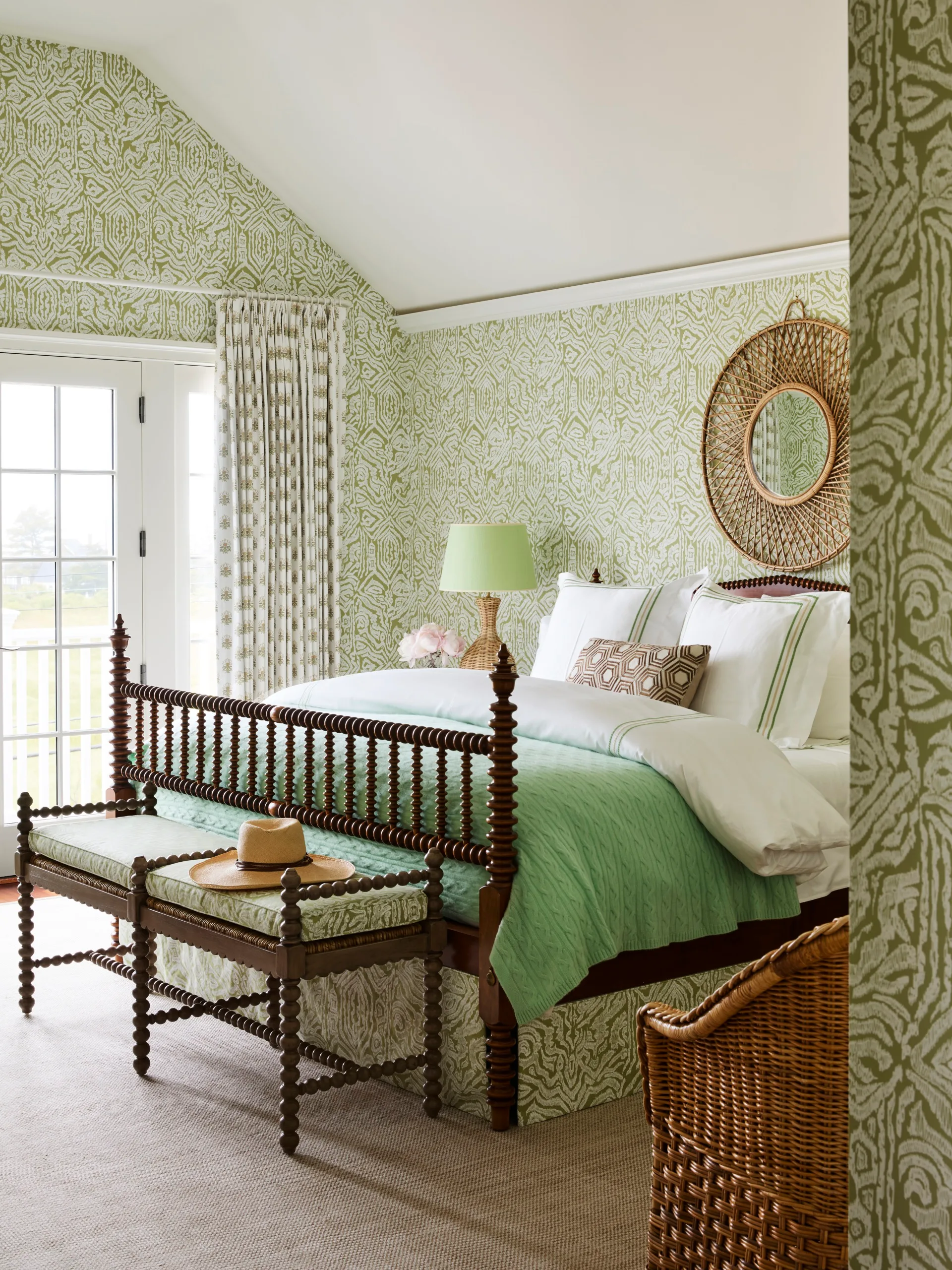 Elegant bedroom with green patterned wallpaper and bedding, wicker chair, and a sunburst mirror.