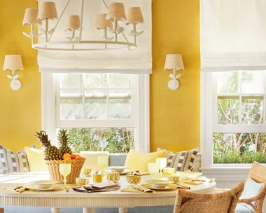 Bright dining room with yellow walls, wicker chairs, a white table set with fruit and dishes, and a large chandelier.