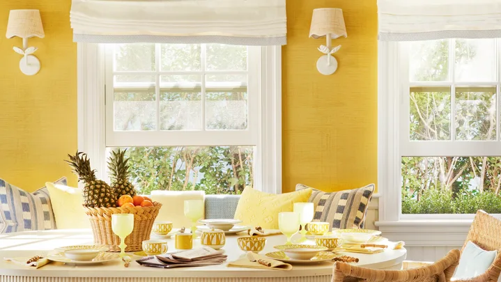Bright dining room with yellow walls, wicker chairs, a white table set with fruit and dishes, and a large chandelier.