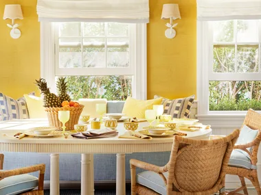 Bright dining room with yellow walls, wicker chairs, a white table set with fruit and dishes, and a large chandelier.