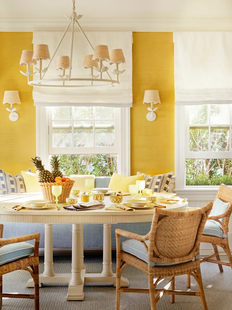 Bright dining room with yellow walls, wicker chairs, a white table set with fruit and dishes, and a large chandelier.
