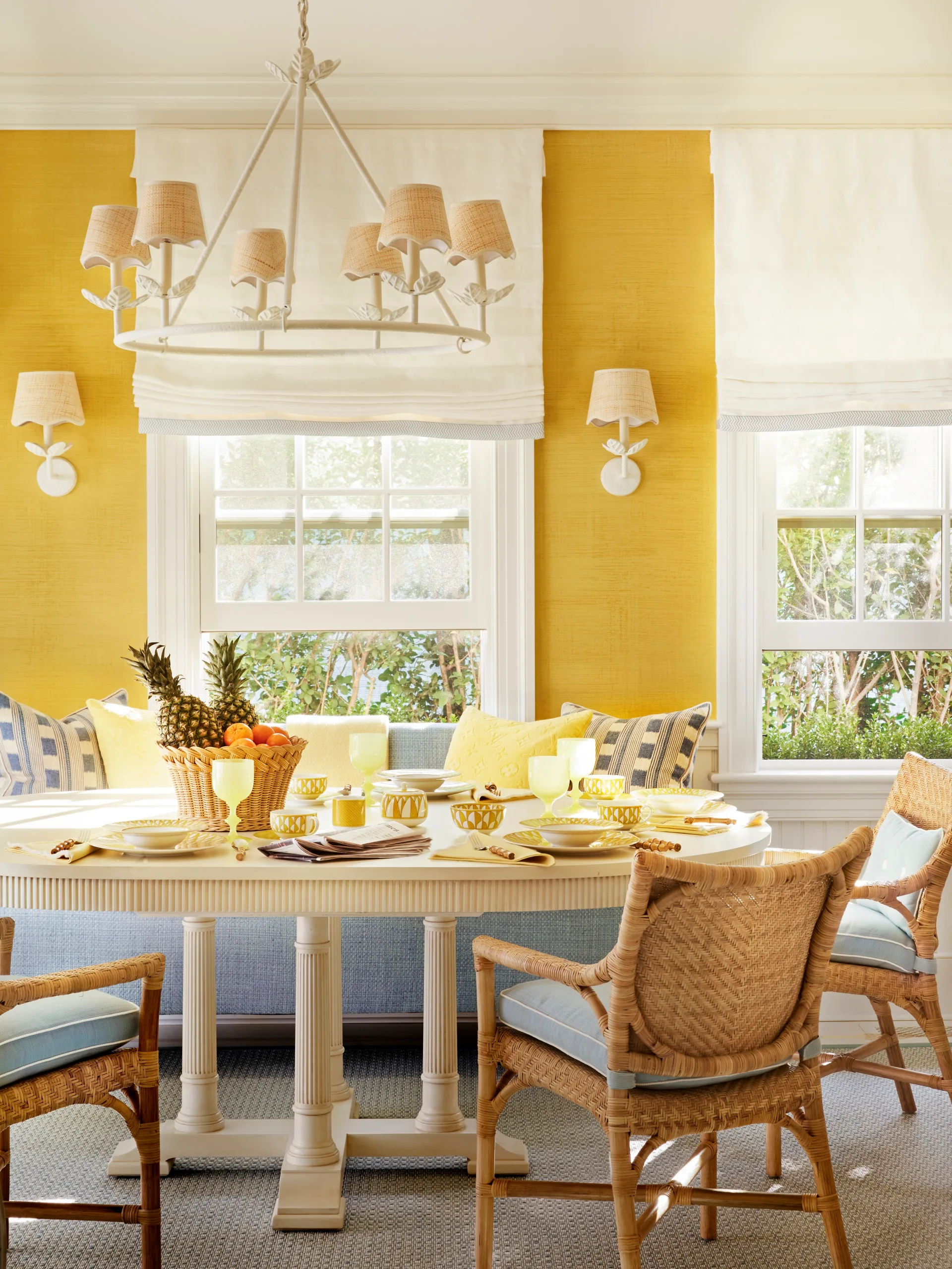 Bright dining room with yellow walls, wicker chairs, a white table set with fruit and dishes, and a large chandelier.