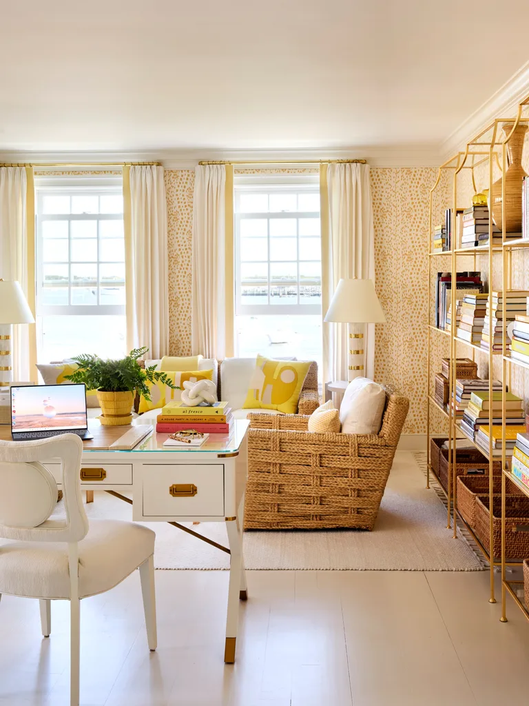 Bright living room with wicker furniture, bookshelves, a white desk, and large windows with light curtains.